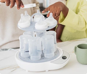 Parent loading baby bottles into a Snookums electric steam steriliser while another parent holds their baby in a modern kitchen.