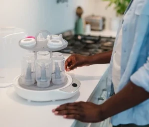 Parent using a Snookums electric steam steriliser to sterilise baby bottles on a kitchen countertop before feeding time.