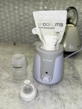 Snookums Electric Bottle Warmer heating a breast milk storage bag with baby bottle and teat beside it